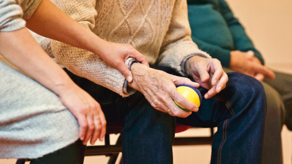 Pflegerin hält behutsam die Hand eines Seniors, der einen Anti-Stress-Ball drückt — ruhige Aktivierungsübung